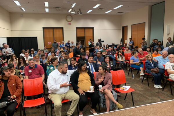 Photo: A crowd of people sit in rows in a large room at the Brownsville Public Library chatting amongst themselves