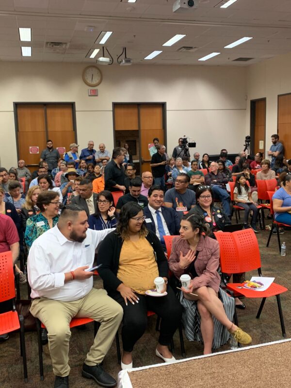 Photo: A crowd of people sit in rows in a large room at the Brownsville Public Library chatting amongst themselves