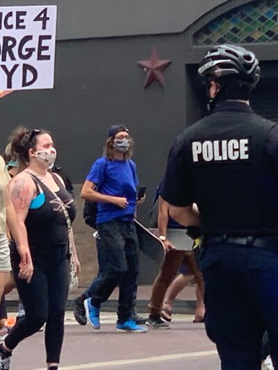 Photo: A police officer looks on as a crowd of people march past at a downtown Houston intersection. In focus is a person holding up a sign that says 'Justice 4 George Floyd.'