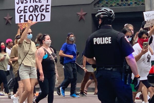 Photo: A police officer looks on as a crowd of people march past at a downtown Houston intersection. In focus is a person holding up a sign that says 'Justice 4 George Floyd.'
