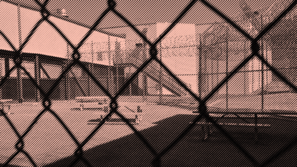 Image: A stylized photo shows an outdoor area of a detention center with some benches and tables. It is surrounded by barbed wire and concertina wire. The camera's view is partially obscured by the barbed wire fencing.