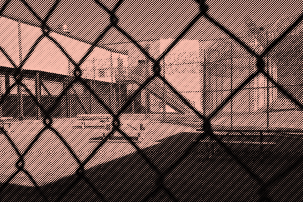 Image: A stylized photo shows an outdoor area of a detention center with some benches and tables. It is surrounded by barbed wire and concertina wire. The camera's view is partially obscured by the barbed wire fencing.