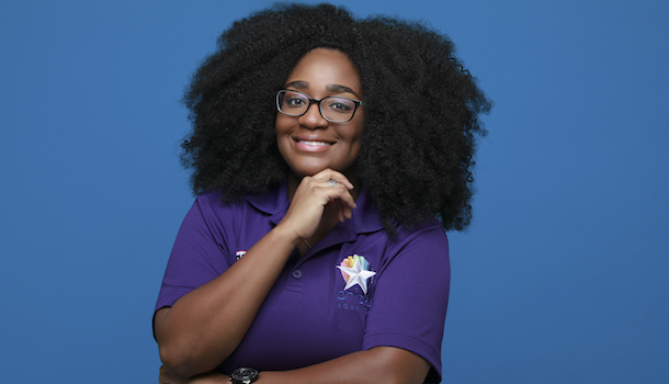 Photo: Lo Roberts poses for a photo in front of a solid background, her hand resting on her chin. She wears a polo with the Pride Houston logo embroidered on it.