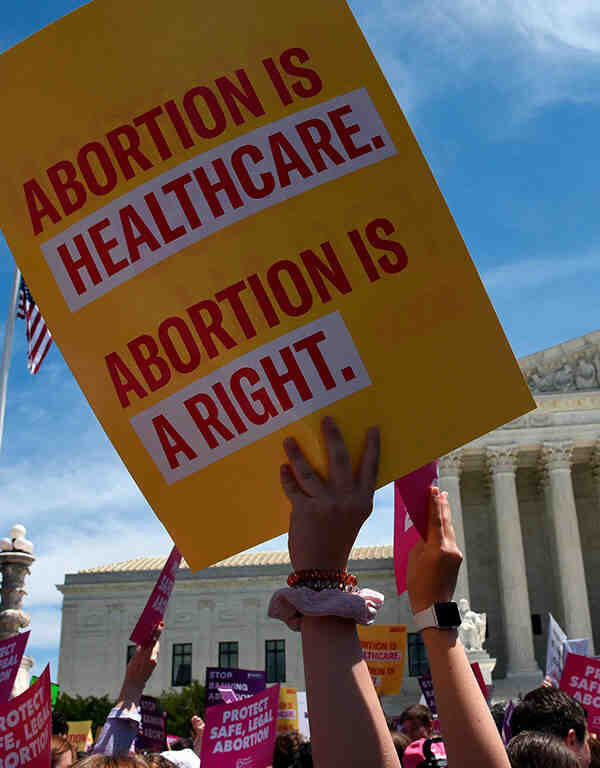 Image: A photo shows the US Supreme Courthouse in the background. Angled upwards, the camera captures the heads and arms of a large crowd of people. Directly in focus is an arm raising a sign that says "Abortion is healthcare. Abortion is a right."
