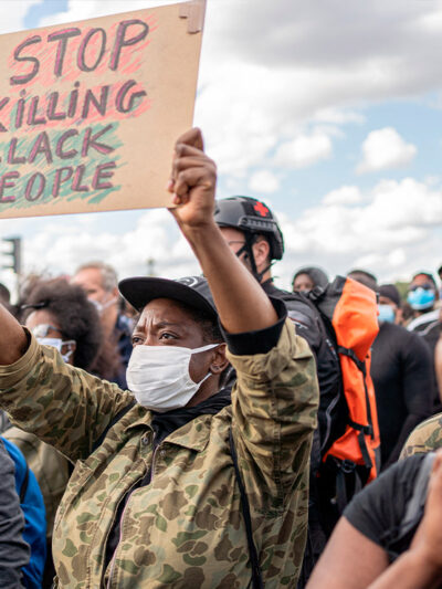 Photo: A crowd of people is gathered and faces one direction. The camera focuses on one African-American person wearing a mask and holding up a sign with handwritten letters that says, "Stop Killing Black People."