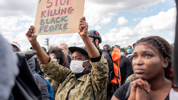Photo: A crowd of people is gathered and faces one direction. The camera focuses on one African-American person wearing a mask and holding up a sign with handwritten letters that says, "Stop Killing Black People."