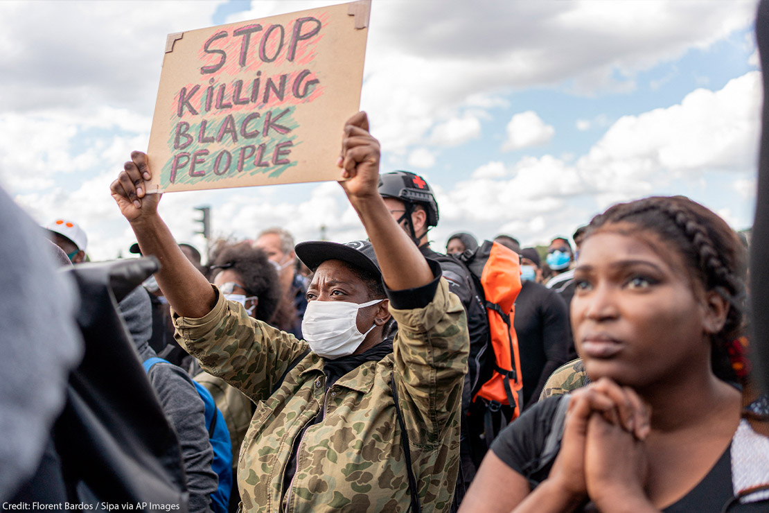 Photo: A crowd of people is gathered and faces one direction. The camera focuses on one African-American person wearing a mask and holding up a sign with handwritten letters that says, "Stop Killing Black People."