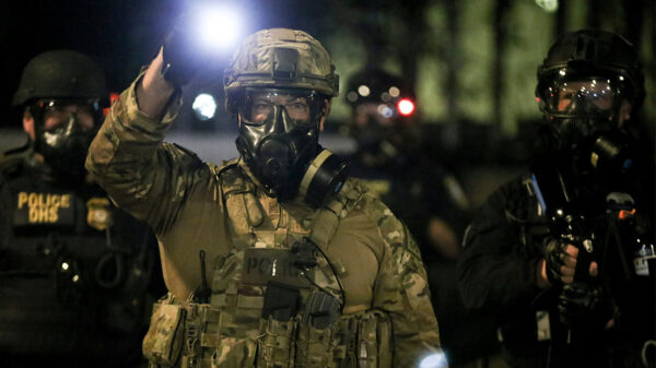 A group of militarized federal agents with an agent in the center shining a bright flashlight toward the viewer.