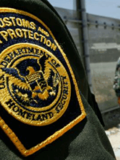 Photo: A Customs and Border Protection patch is worn on a green uniform. The CBP officer's arm takes us most of the photo, a fence and another CBP officer stand in the background.
