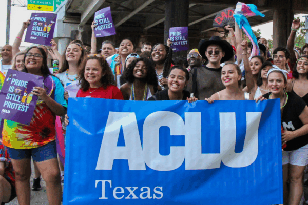 Photo: A large crowd poses for a photo behind a large ACLU of Texas banner. They are assembled under a Houston overpass, many holding signs that say 'Pride is still Protest."