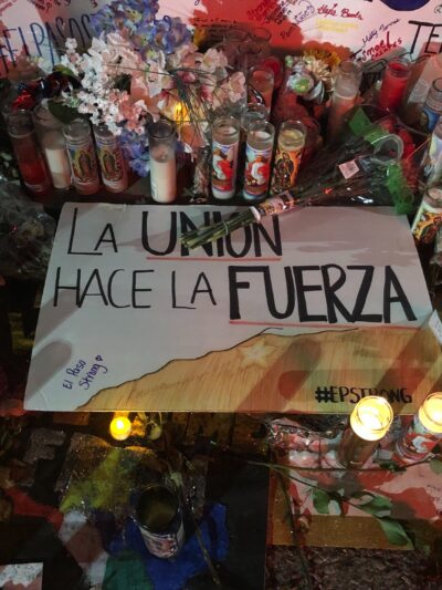 Photo: Handwritten signs, candles, and flowers adorn a section of a street. Pictured at center is a sign that says "La union hace la fuerza."