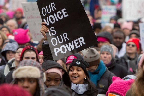 Photo: A crowd of people stand together, many wearing pink beanies or holding signs. Centered is a woman holding up a printed sign that reads:"Our voice is our power."