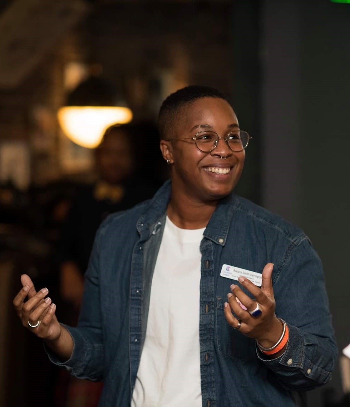 Photo: Avatara Smith-Carrington is grinning and gesturing as they look off camera. They were a nametag