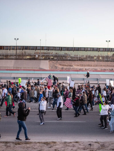 Photo: A large crowd streams by a a freeway and border fencing. They carry signs and american flags, walking in a general procession.