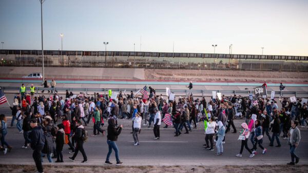 Photo: A large crowd streams by a a freeway and border fencing. They carry signs and american flags, walking in a general procession.