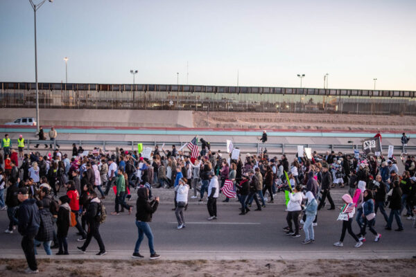 Photo: A large crowd streams by a a freeway and border fencing. They carry signs and american flags, walking in a general procession.