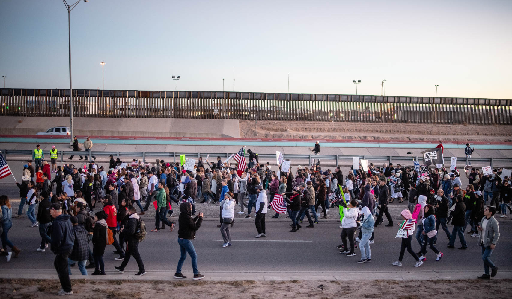 Photo: A large crowd streams by a a freeway and border fencing. They carry signs and american flags, walking in a general procession.