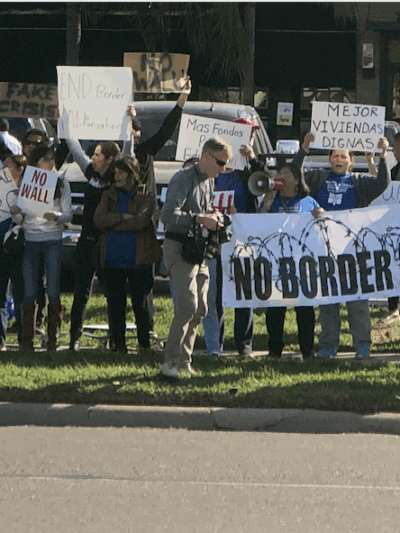 Photo: A crowd of people stand along a street in the Rio Grande Valley. Behind them is a restaurant and other businesses. They hold signs, one of which says "No Border Wall."