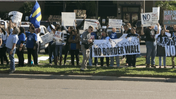 Photo: A crowd of people stand along a street in the Rio Grande Valley. Behind them is a restaurant and other businesses. They hold signs, one of which says "No Border Wall."