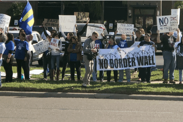 Photo: A crowd of people stand along a street in the Rio Grande Valley. Behind them is a restaurant and other businesses. They hold signs, one of which says "No Border Wall."