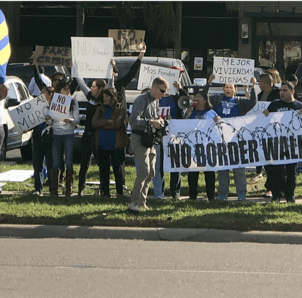 Photo: A crowd of people stand along a street in the Rio Grande Valley. Behind them is a restaurant and other businesses. They hold signs, one of which says "No Border Wall."