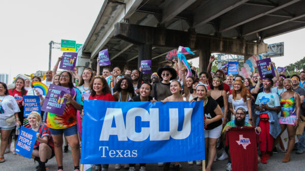 Photo: A large crowd in colorful clothing gathers under an overpass. Many hold signs that say "Pride is still protest" or "Trans people belong." A few in the foreground hold a large vynl banner that says "ACLU of Texas"