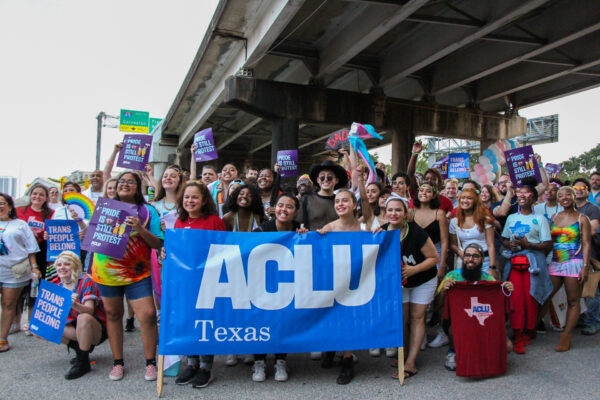 Photo: A large crowd in colorful clothing gathers under an overpass. Many hold signs that say "Pride is still protest" or "Trans people belong." A few in the foreground hold a large vynl banner that says "ACLU of Texas"