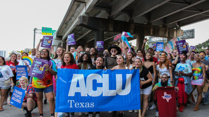 Photo: A large crowd in colorful clothing gathers under an overpass. Many hold signs that say "Pride is still protest" or "Trans people belong." A few in the foreground hold a large vynl banner that says "ACLU of Texas"