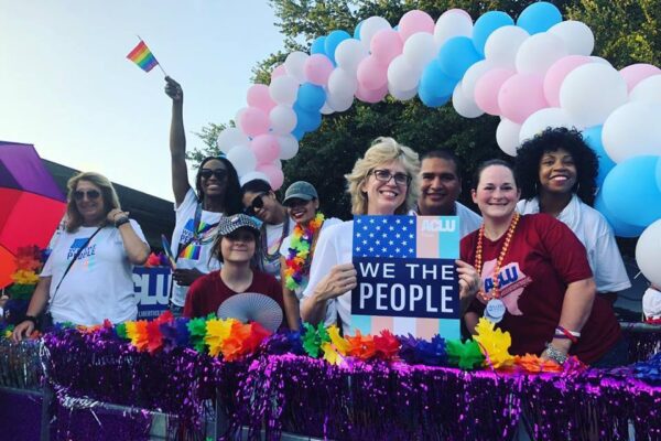Photo: People pose for a photo on a float decorated with rainbow flower garlands and shimmery streamers. Arching behind them is a balloon sculpture in alternating trans pride colors.