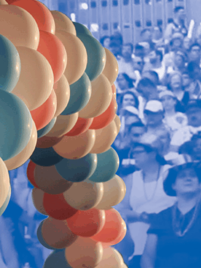 Image: An image of balloon arch with alternating trans pride colors overlaps a stylized image of a crowd of people gathered.