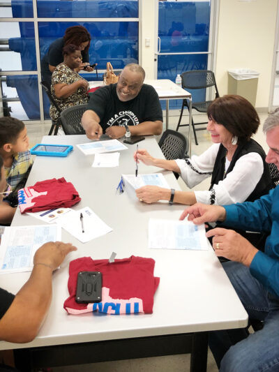 Image: People of various ages gather around a table with paper and ACLU of Texas T shirts on it. A Black man towards the end of the table gestures as he speaks.