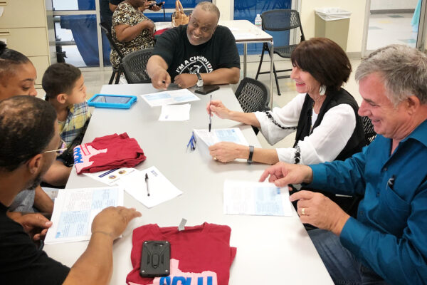 Image: People of various ages gather around a table with paper and ACLU of Texas T shirts on it. A Black man towards the end of the table gestures as he speaks.