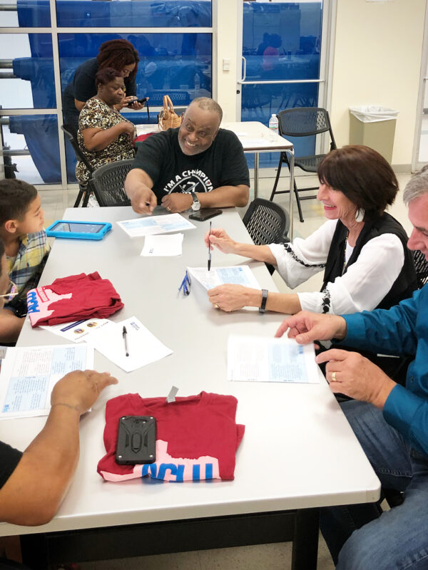 Image: People of various ages gather around a table with paper and ACLU of Texas T shirts on it. A Black man towards the end of the table gestures as he speaks.