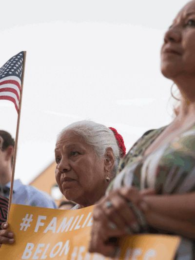 Image: Three women look on towards an unpictured speaker. They hold signs that say with the hashtag #FamiliesBelongTogether. One holds a U.S. flag up with her sign.