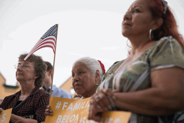 Image: Three women look on towards an unpictured speaker. They hold signs that say with the hashtag #FamiliesBelongTogether. One holds a U.S. flag up with her sign.