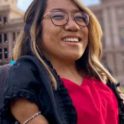 A disabled Mexican-American woman with medium-length hair. She is smiling to the camera, has glasses, and is wearing a black blazer with a pink blouse.