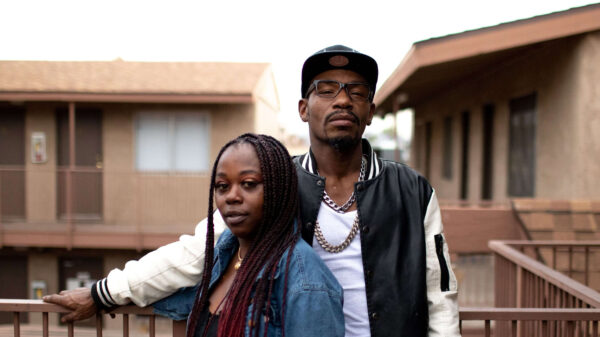 A young Black couple stand side by side on a balcony posing for the camera