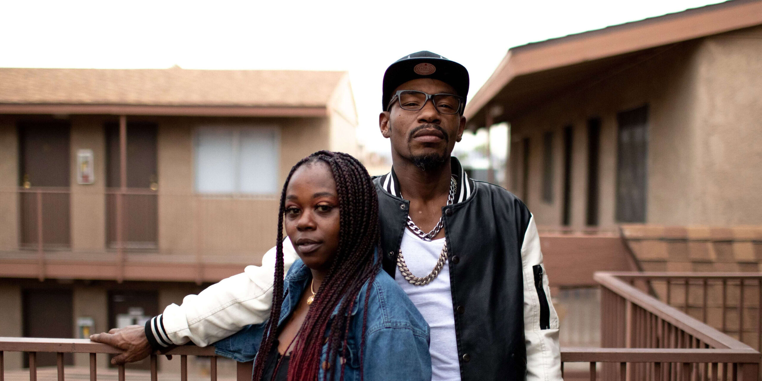 A young Black couple stand side by side on a balcony posing for the camera