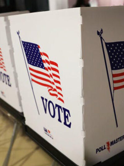 Photo: People lean into individual polling booths arranged in a line in a school gym. Their faces are obscured by divders between booths.