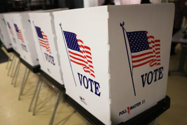 Photo: People lean into individual polling booths arranged in a line in a school gym. Their faces are obscured by divders between booths.
