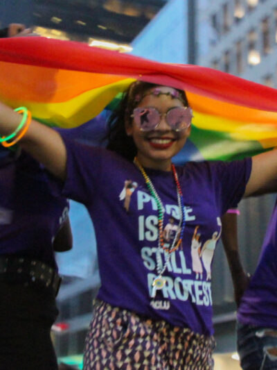 Smiling woman standing with rainbow flag above her head wearing t-shirt that says Pride is Still Protest
