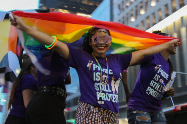 Smiling woman standing with rainbow flag above her head wearing t-shirt that says Pride is Still Protest