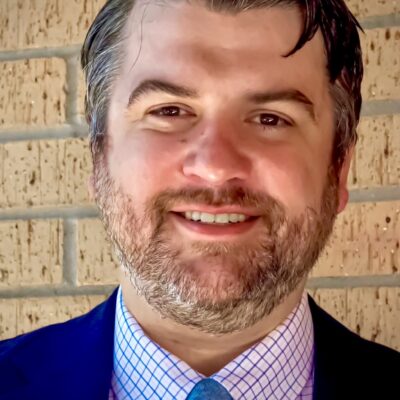 Person with short brown hair, plaid collared shirt, blue tie and dress coat