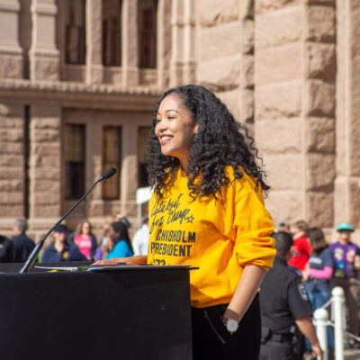Photo of person standing at a podium smiling with curly dark hair, brown skin, wearing a yellow sweatshirt and black pants