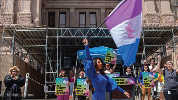 A drag queen raises a flag in front of supporters holding signs.