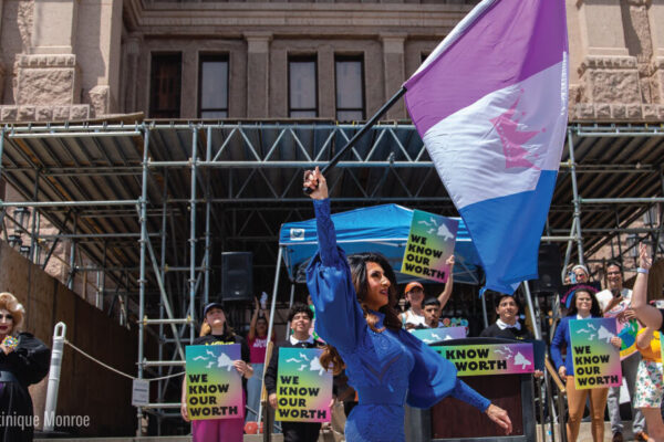 A drag queen raises a flag in front of supporters holding signs.