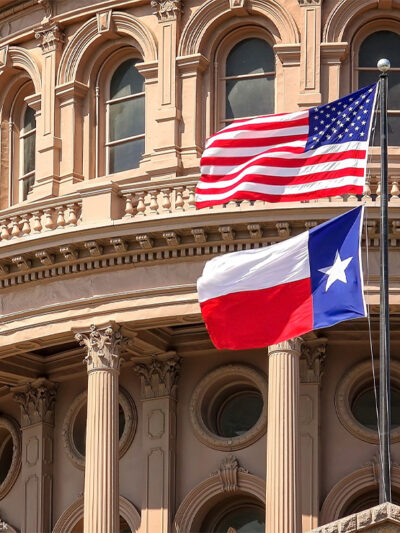 American and Texas state flags flying on the dome of the Texas State Capitol building.