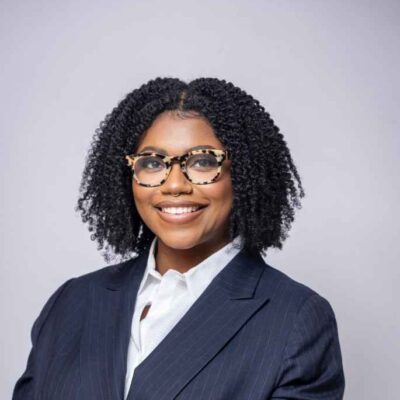 Professional headshot taken from the shoulders up of a Black person wearing a suit and tortoise-shell glasses. They have shoulder-length curly hair and are smiling brightly toward the camera.