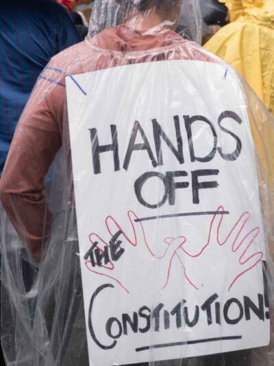 Photo of a person at a protest, taken from behind, wearing a protest sign from their back that reads "Hands off the Constitution"
