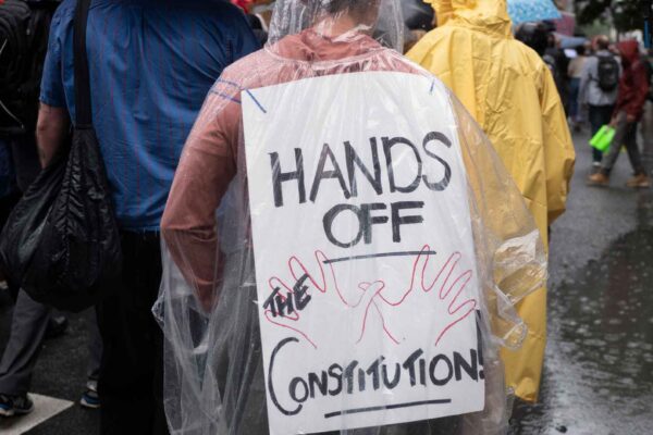 Photo of a person at a protest, taken from behind, wearing a protest sign from their back that reads "Hands off the Constitution"